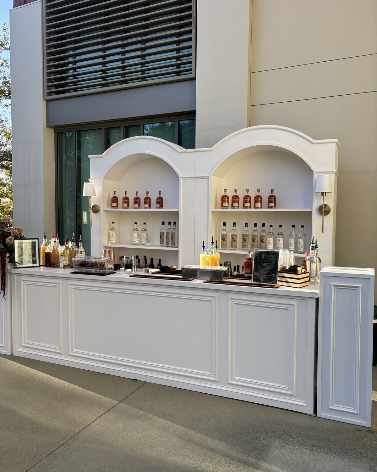 A white arched back bar with lit sconces and stocked shelves, set and ready before the first guest arrives